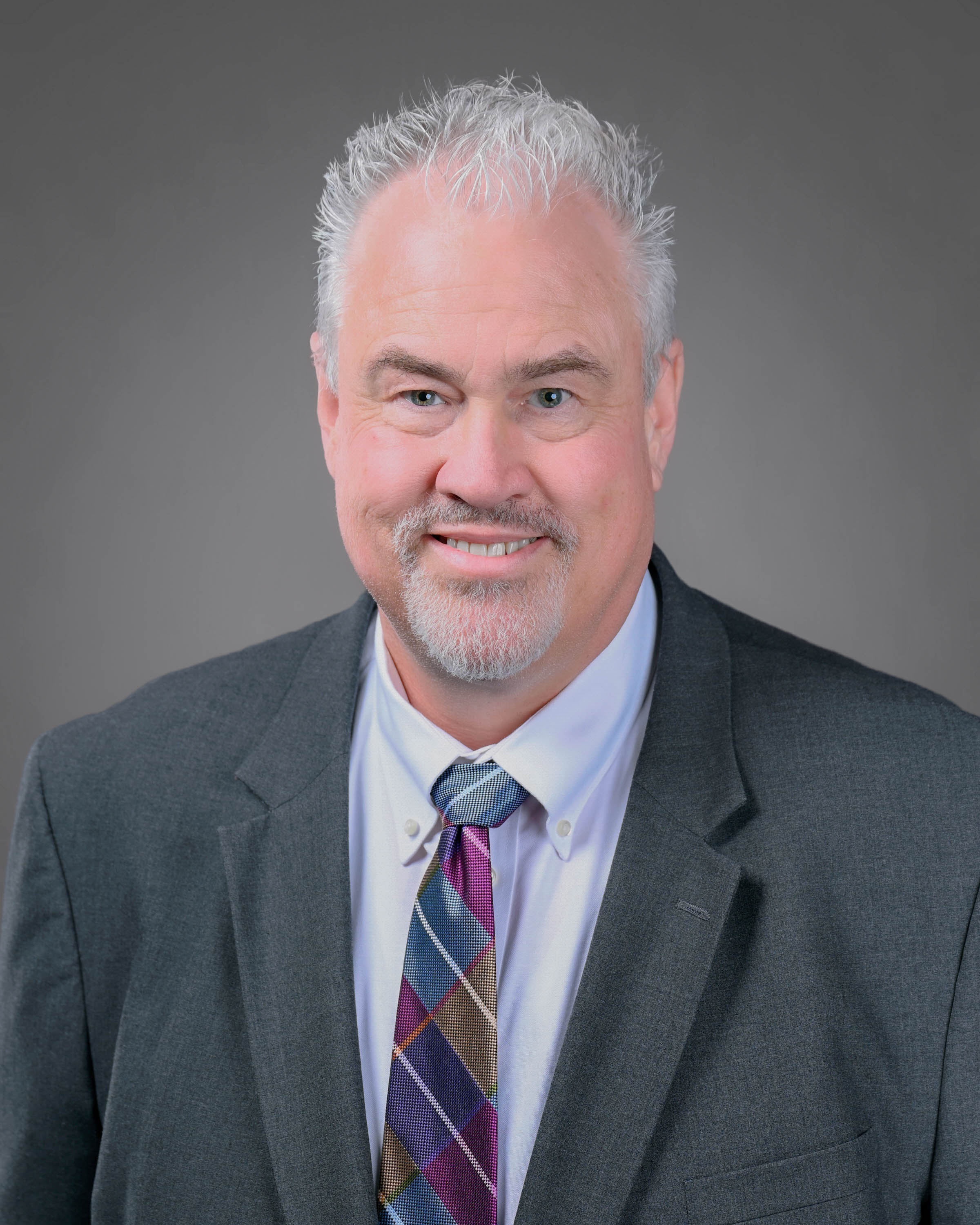 Professional headshot of a smiling man with short white hair and a goatee, wearing a dark gray suit, white dress shirt, and multicolored patterned tie against a gray studio background.